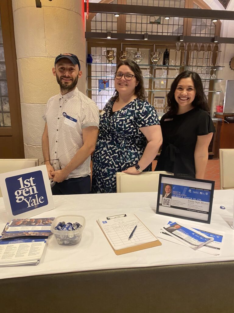 Three people smiling and standing behind a table with 1stGenYale promotional buttons and flyers