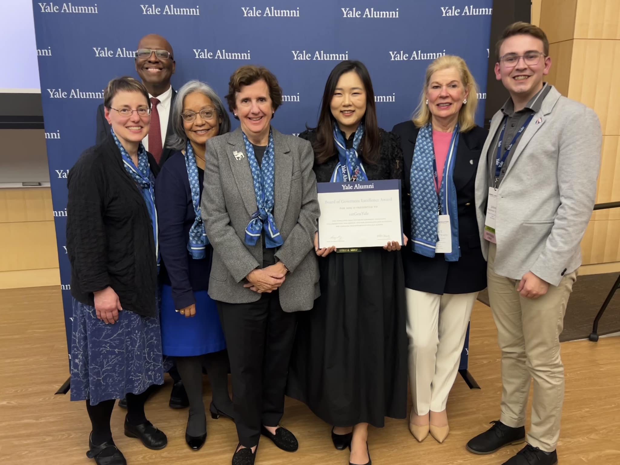 Seven alums stand hold an award certificate and stand in front of a blue banner reading Yale Alumni