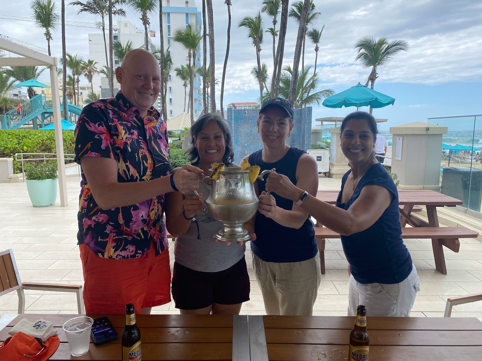 Four alums stand holding a Mory's cup with palm trees and a beach in the background