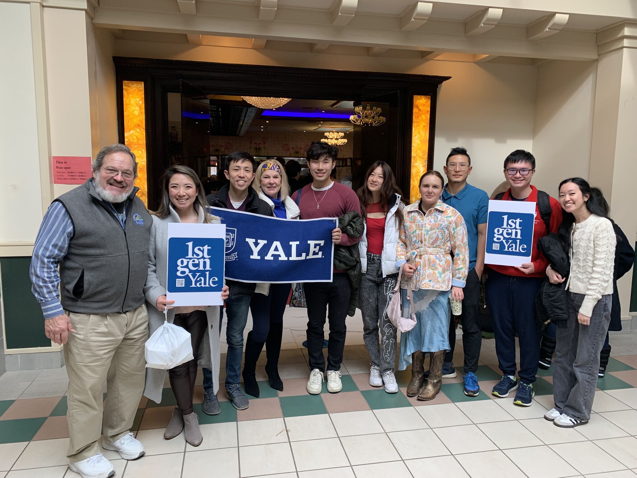 Ten alums stand outside a restaurant and hold Yale and 1stGenYale signs