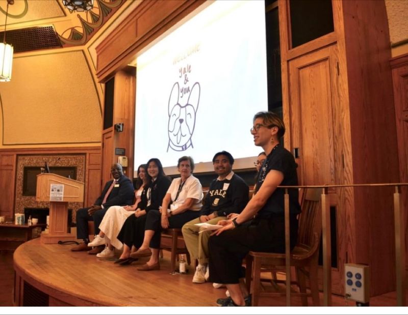 Seven people sit on a stage in front of a screen with a drawing of a bulldog and the words Yale & You