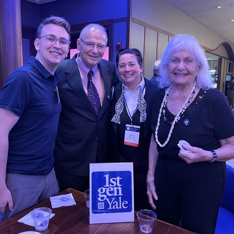 Several alums stand near a table with a 1stGenYale sign