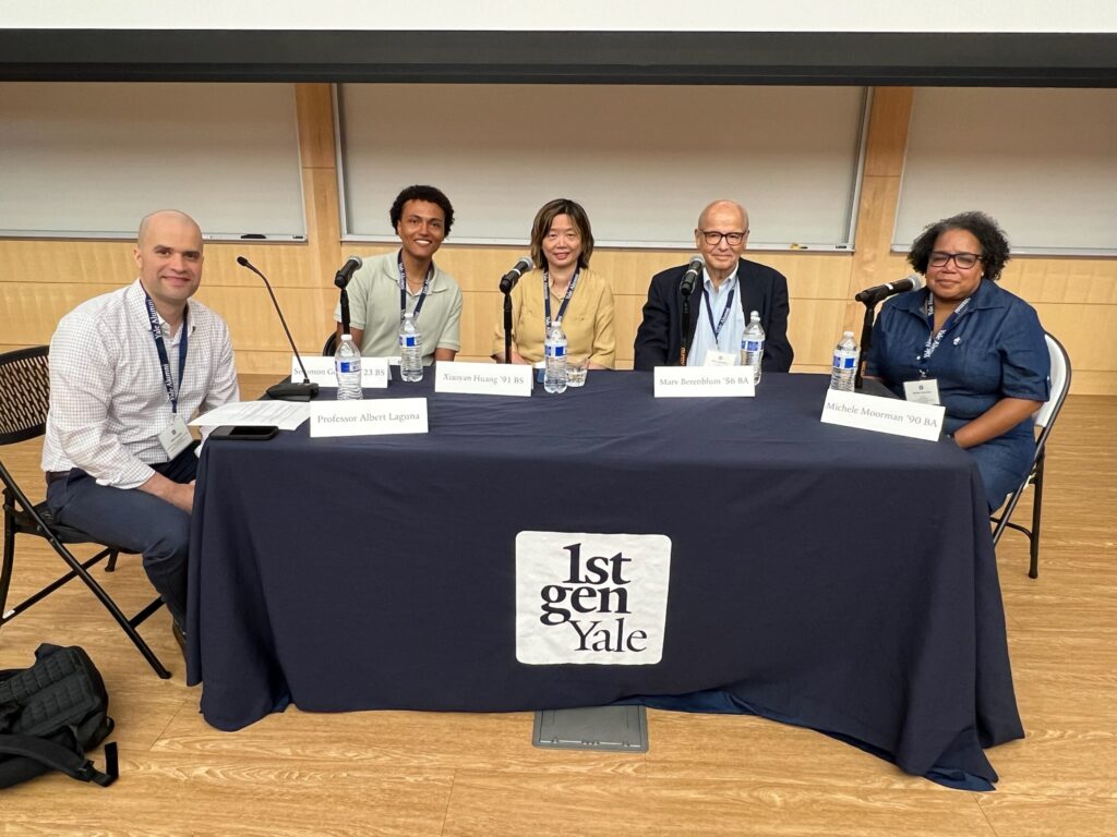 Five people sit around a table on a stage. The table has a 1stGenYale banner on it, and there are microphones and name plates in front of the people