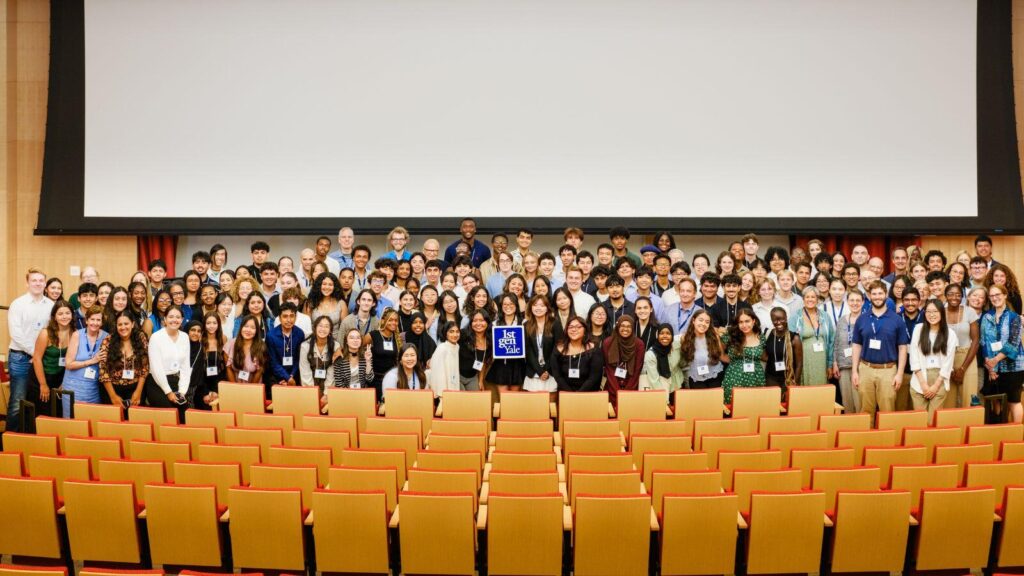 More than a hundred people stand in several rows across the front stage of a large auditorium